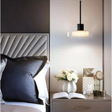A glass disc pendant lit above a nightstand beside a tufted headboard and an open book
