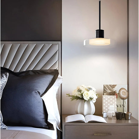 A glass disc pendant lit above a nightstand beside a tufted headboard and an open book