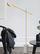 The gold lamp reaching over a chair with a coat on it beside a plant and a table