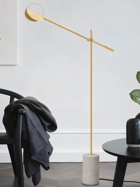 The gold lamp reaching over a chair with a coat on it beside a plant and a table