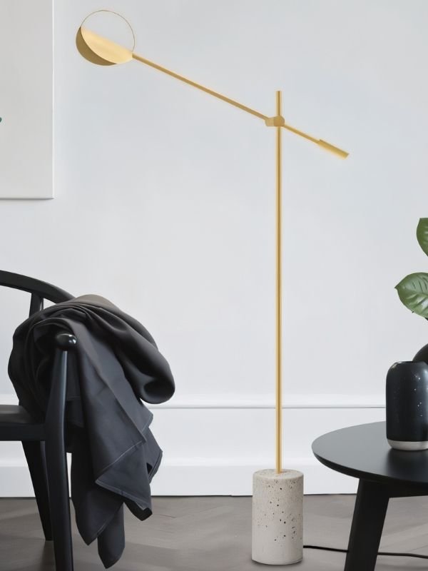 The gold lamp reaching over a chair with a coat on it beside a plant and a table