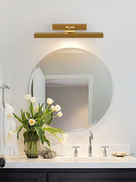Gold bar above a round mirror in a bathroom, flowers beside the basin