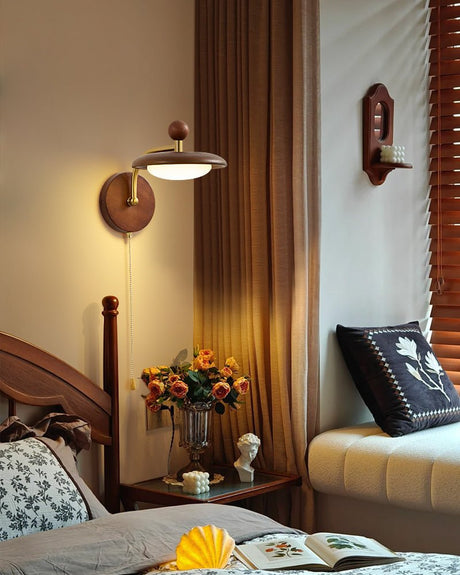 Sconce lit above a bedside table with blossom in a jug beside a spindle bed