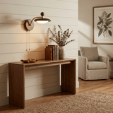 Sconce lit above a walnut console beside an armchair on a woven rug
