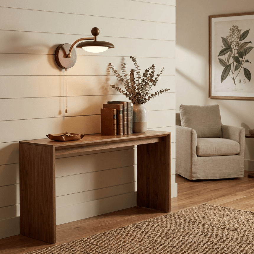 Sconce lit above a walnut console beside an armchair on a woven rug