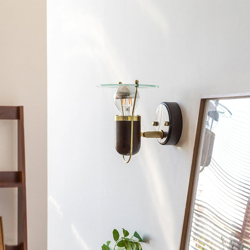 Sconce lit beside a leaning mirror and a plant against a white wall