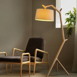 The lit lamp leaning over two wooden armchairs beside a plant and a window