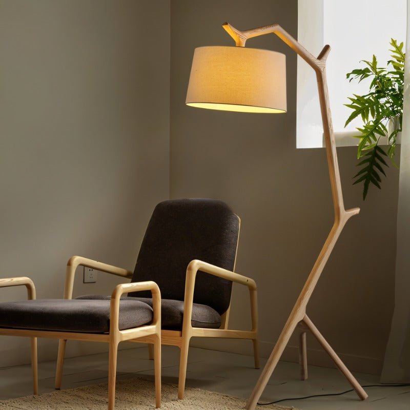 The lit lamp leaning over two wooden armchairs beside a plant and a window