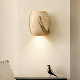 The travertine urn lit above a black floating shelf with a bird figure, a woven chair below