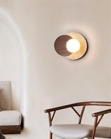 Sconce lit on a plaster wall beside a low wood chair and a cushion