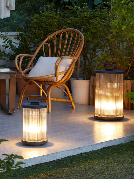 Two lanterns lit on paving at the edge of a lawn beside a woven chair