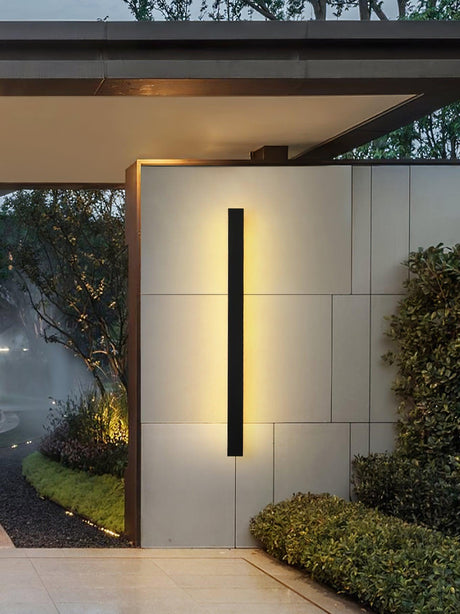 Black linear strip lit on a pale entry wall between a hedge and a covered path