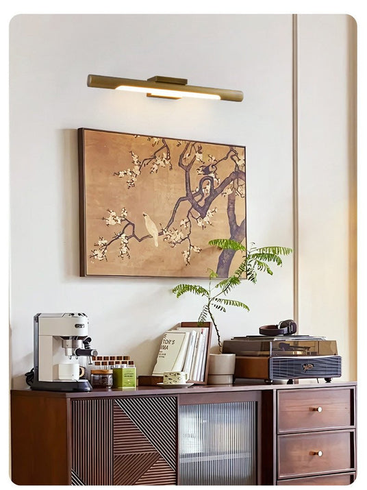 Brass tube lit over a framed painting above a wood sideboard and plant