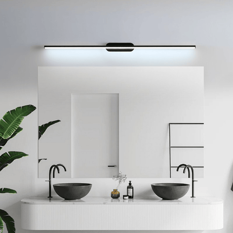 Thin rod lit above a bathroom mirror over two basins beside a large leafy plant