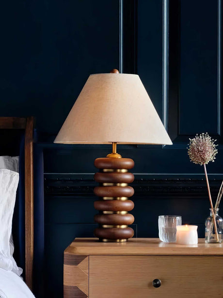 Linen shade table lamp lit on a nightstand beside a bed