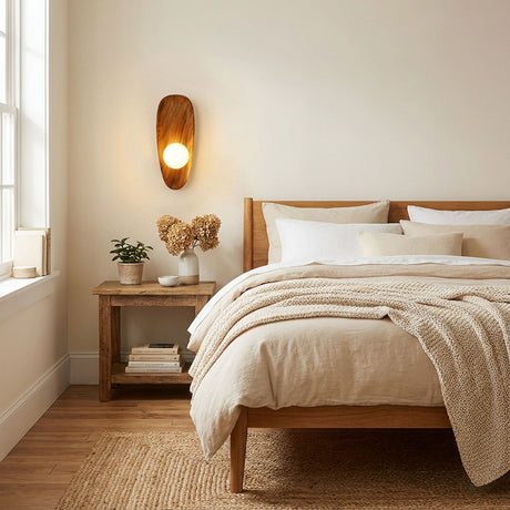 Sconce lit beside a bed above a wood nightstand holding dried flowers