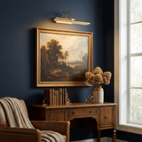 Gold bar lit above a framed landscape on a navy wall over a wood desk and chair