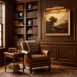 Gold bar lit above a framed painting in a paneled study with a leather armchair