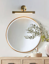 Bronze bar lit above a round mirror on a wood console with a plant and bottles