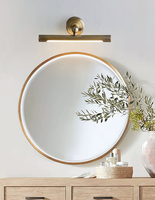 Bronze bar lit above a round mirror on a wood console with a plant and bottles