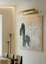 Brass bar lit over a framed artwork on a wall beside dining chairs