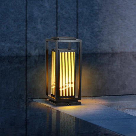 The lantern lit on paving beside still water with planting behind