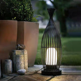 The lantern lit on paving beside tall planters and a low stone