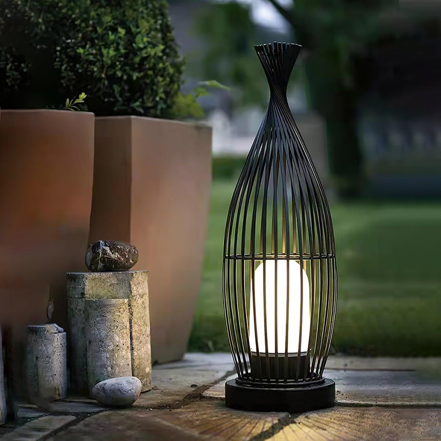 The lantern lit on paving beside tall planters and a low stone