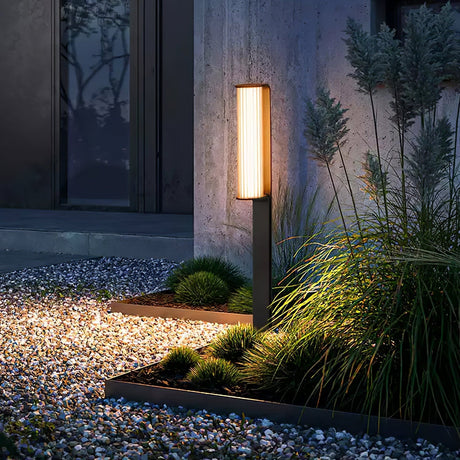 One arm lit at a gravel edge beside grasses and a lit house wall