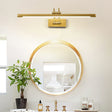 Brass bar lit above a round gold mirror on a vanity with a plant and glass bottles