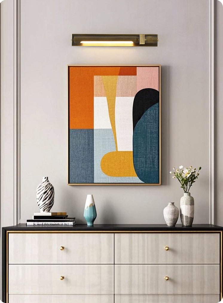 Bronze bar lit over a framed canvas above a pale wood sideboard