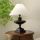Black oversized table lamp lit on a round wooden table beside foliage