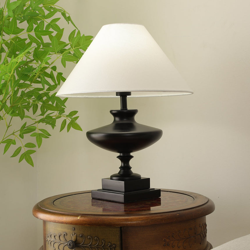 Black oversized table lamp lit on a round wooden table beside foliage