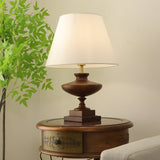 Walnut oversized table lamp on a round table with foliage behind it