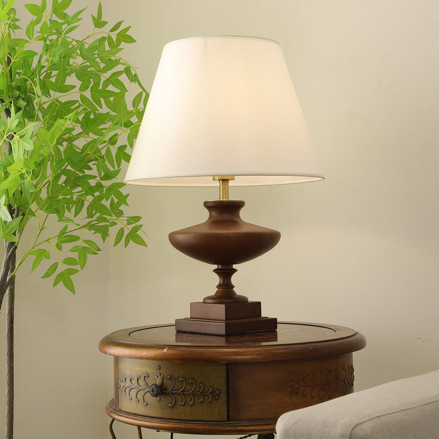 Walnut oversized table lamp on a round table with foliage behind it