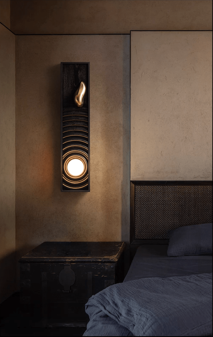 Sconce lit on a wall above a bed with pale bedding in a quiet corner