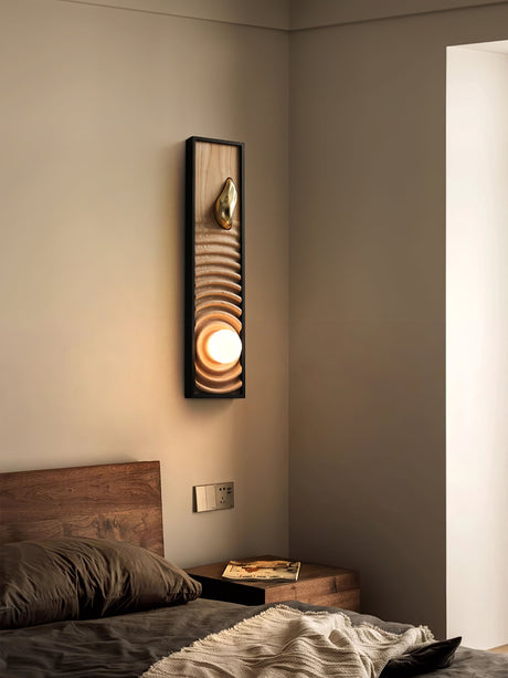 Sconce lit on a bedroom wall in a corner beside a bed with folded linen