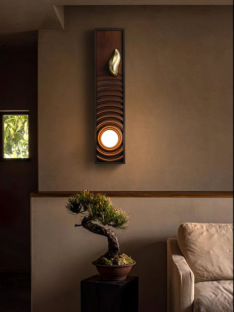 Sconce lit above a bonsai on a stand beside a pale armchair and a window