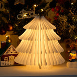 White paper tree lamp lit on a table in front of a decorated tree