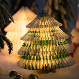 Green paper tree lamp lit beside small baubles and pine branches
