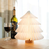 White paper tree lamp lit on a table beside a bottle and two glasses