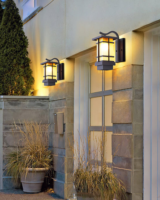 Two lanterns lit on a courtyard wall beside a glass door, potted grasses below