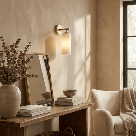 The sconce lit above a desk with a leaning mirror and a stack of books