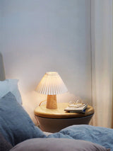Pleated table lamp lit on a round nightstand beside a bed with blue linen