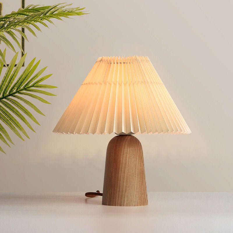 Pleated table lamp lit warm, the folds glowing beside a palm frond