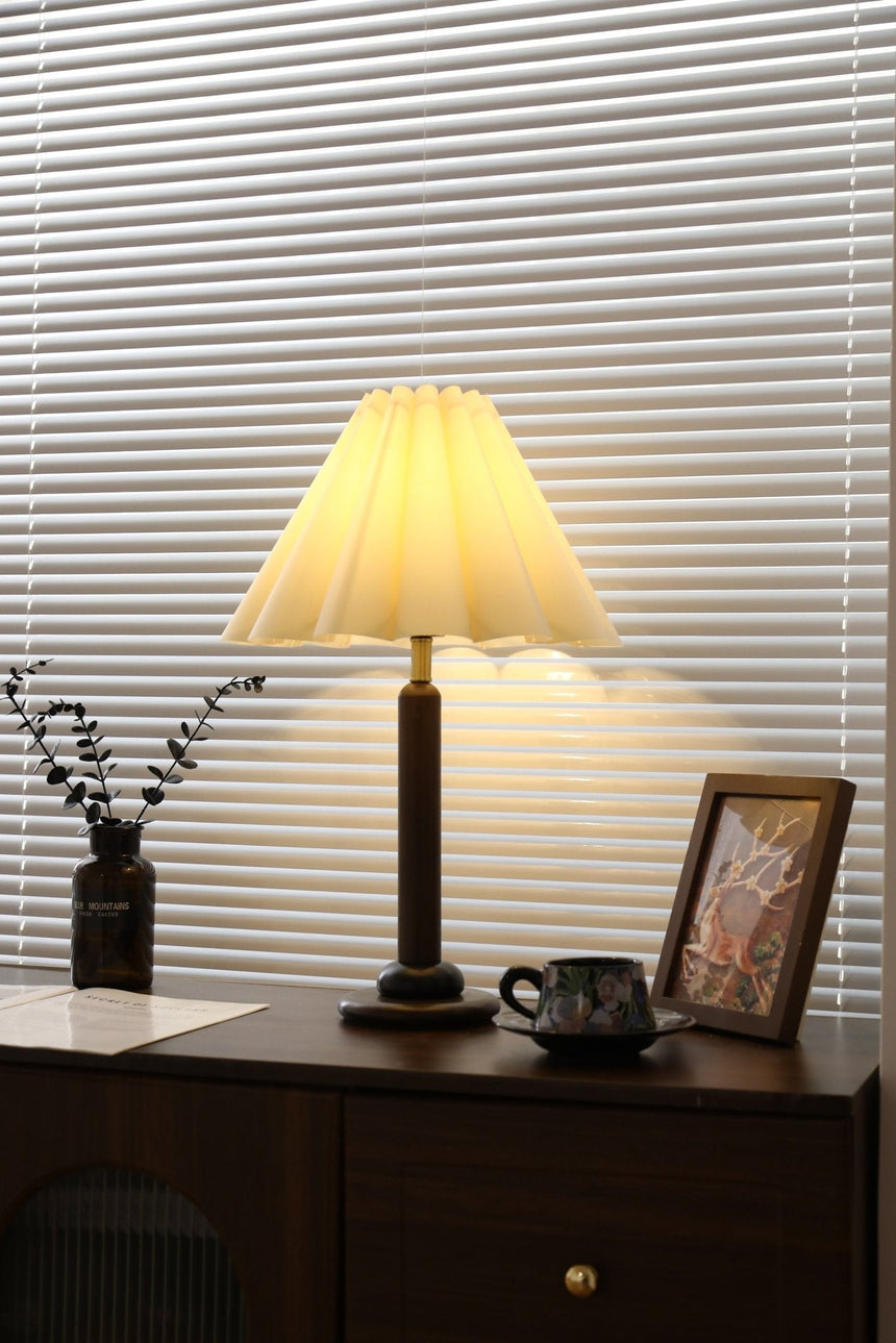 Pleated table lamp lit close, the linen cone and wood column in view