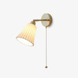 Brass wall sconce with a pleated white ceramic trumpet shade and a pull chain, lit