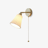 Brass wall sconce with a pleated white ceramic trumpet shade and a pull chain, lit