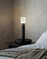 Black portable table lamp lit on a nightstand beside a made bed