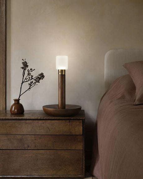 Portable table lamp lit on a dark wood nightstand next to a bed
